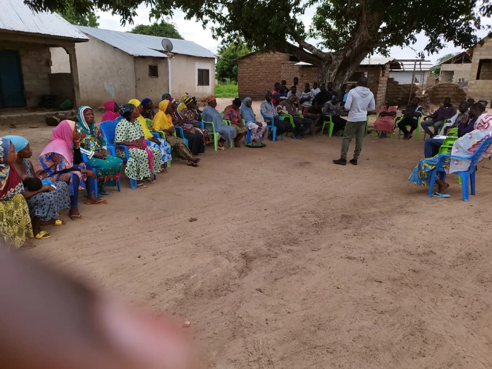 Community training session under a tree; women farmers on chairs, facilitator standing